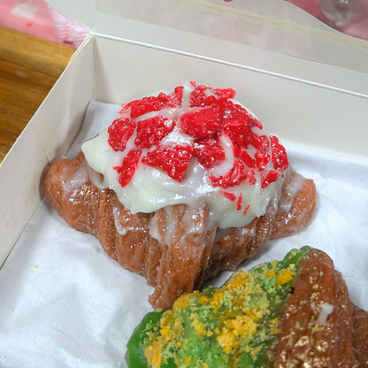 Two decorated pastry candles inside a white cardboard box on white paper. The top pastry is a croissant with white icing and red crumb topping, and the bottom pastry is partially visible with green and yellow topping.