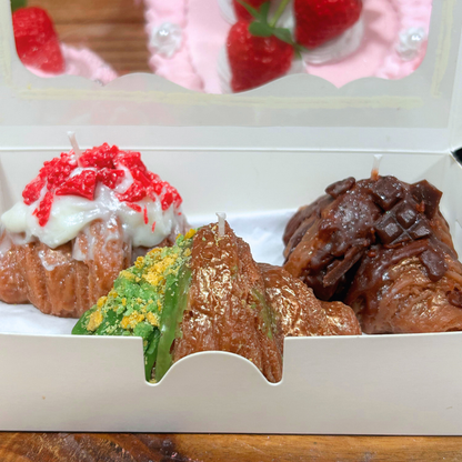 A close-up of a white bakery box containing three assorted candles: a strawberry-topped croissant, a green and yellow crumb-topped pastry, a chocolate-covered pastry.