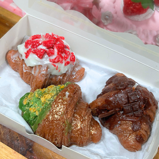 Three decorated croissant-style candles arranged inside a white bakery box on white parchment paper. One pastry has white frosting topped with red crumb pieces, one has green frosting with yellow crumbs, and one is dark chocolate brown with a grid pattern.