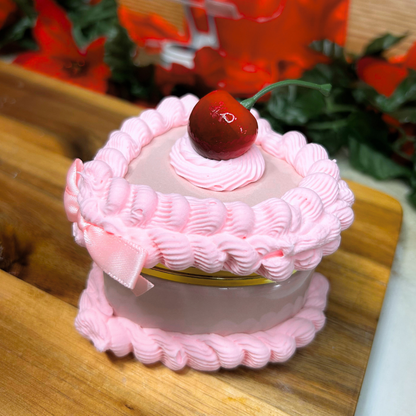 Pink cake-shaped trinket box with a cherry on top and pink bow on a wooden surface