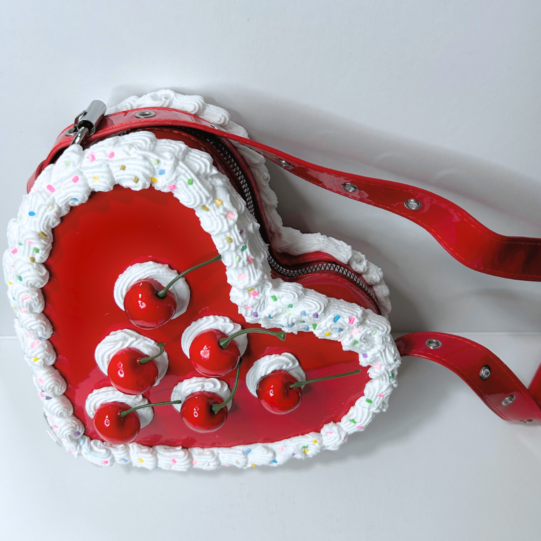 Red heart-shaped cake bag with white frosting, cherries, and colorful sprinkles on a white background