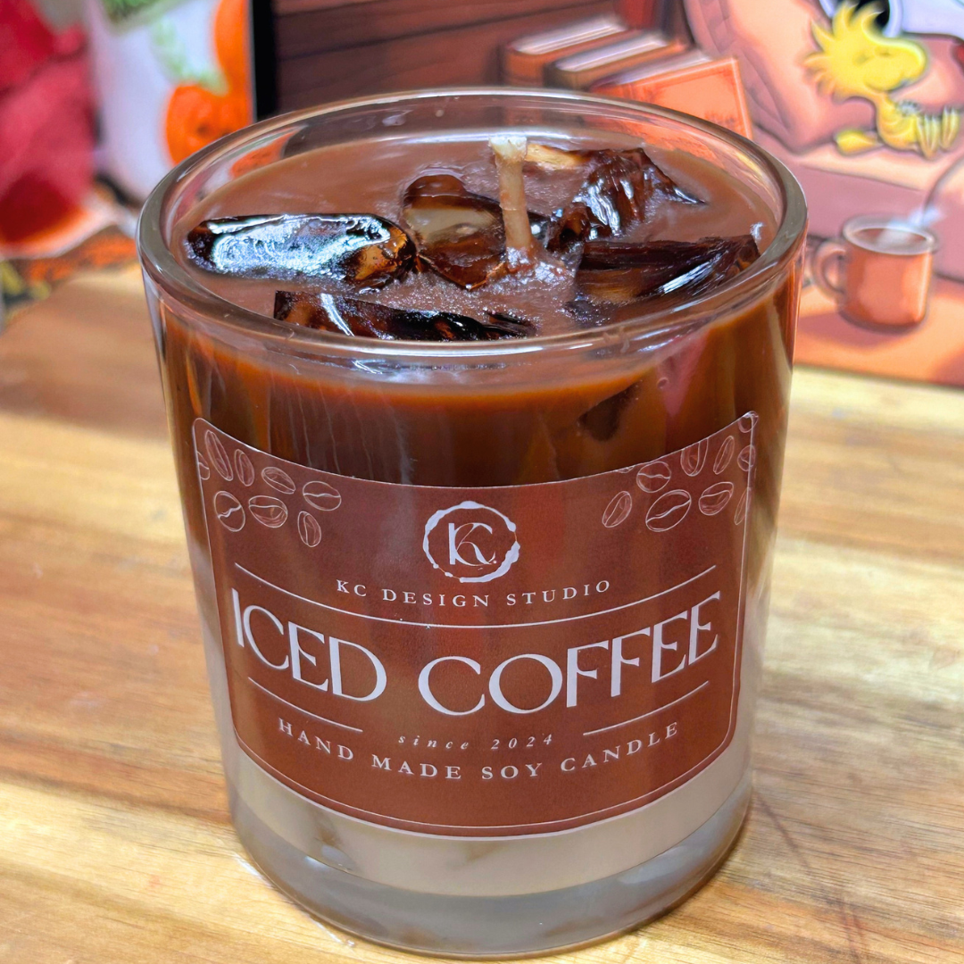 A soy candle in a clear glass vessel with an iced coffee themed label, filled with dark brown wax and ice cubes, sitting on a wooden surface.