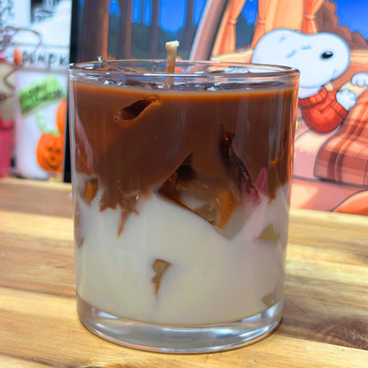 A layered candle in a clear glass jar with a wick, showing a dark brown top layer and a creamy off-white bottom layer, set on a wooden surface with a colorful cartoon backdrop.