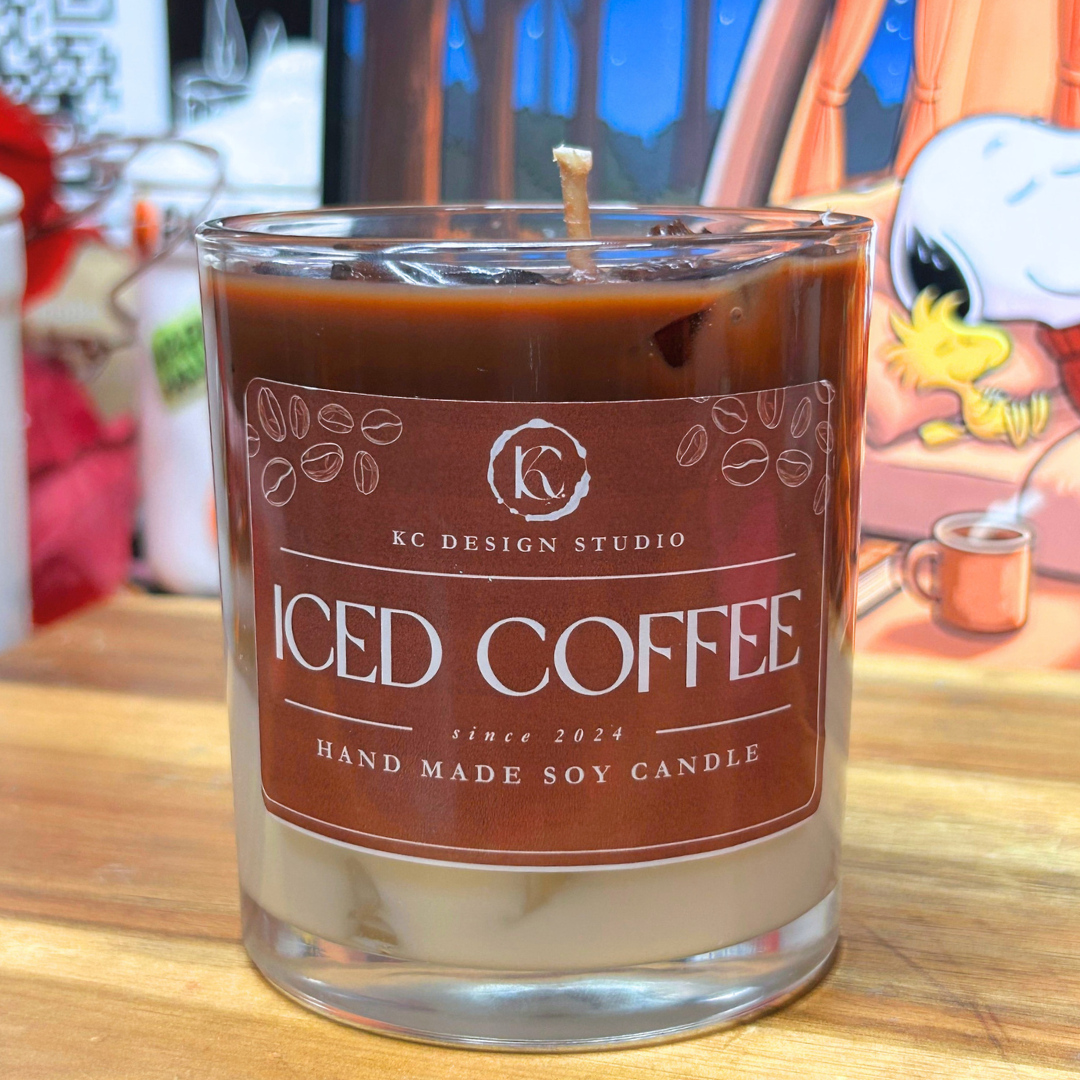 A glass jar candle labeled 'ICED COFFEE' by KC Design Studio, with a brown wax top layer, cream-colored wax bottom layer, and a single wick, set on a wooden surface with a blurred background.