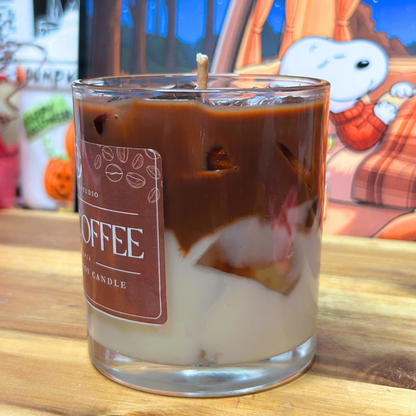 A glass jar candle with a wick and layered design resembling coffee and milk, with a brown label reading 'COFFEE' on a wooden table. A Snoopy-themed backdrop is visible in the background.