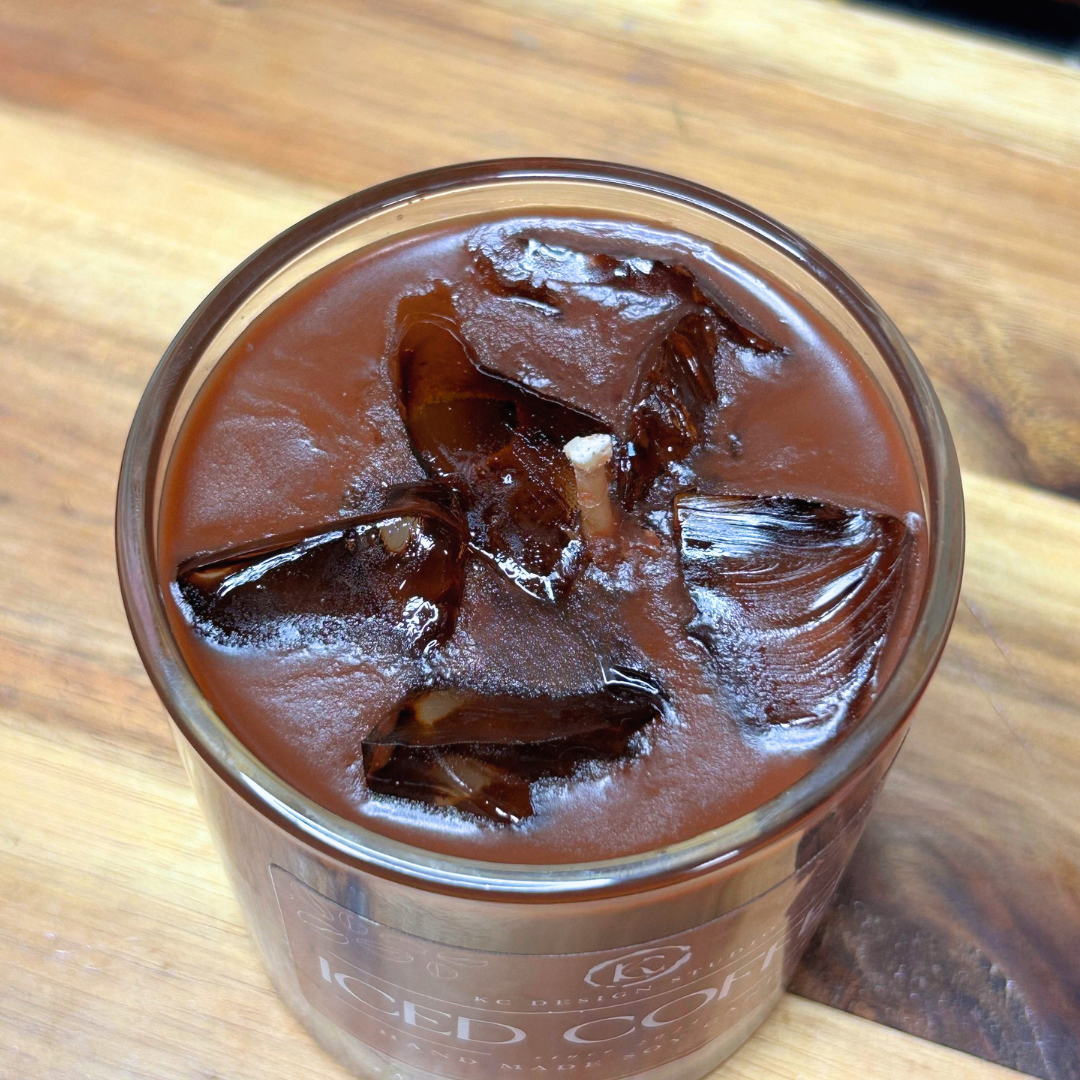 A top-down close-up of a glass jar candle filled with a rich, dark brown liquid and large ice cubes, labeled 'ICED COFFEE', resting on a wooden surface.
