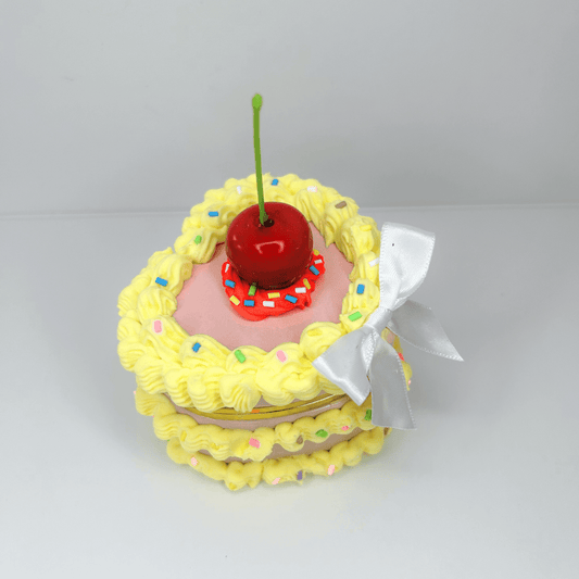 Top view of a yellow heart-shaped mini jewelry box decorated as a cake, featuring colorful sprinkles and a cherry on top.