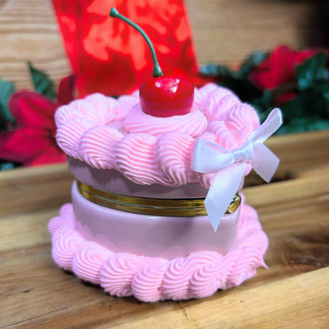 Pink cake-shaped trinket box with a cherry on top and a bow, set against a blurred floral background.