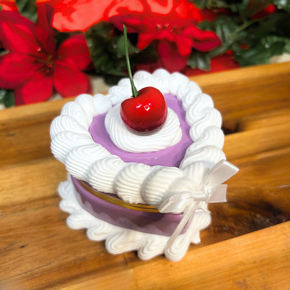 Decorative purple cake-shaped trinket box with a cherry on top on a wooden surface with red flowers in the background.