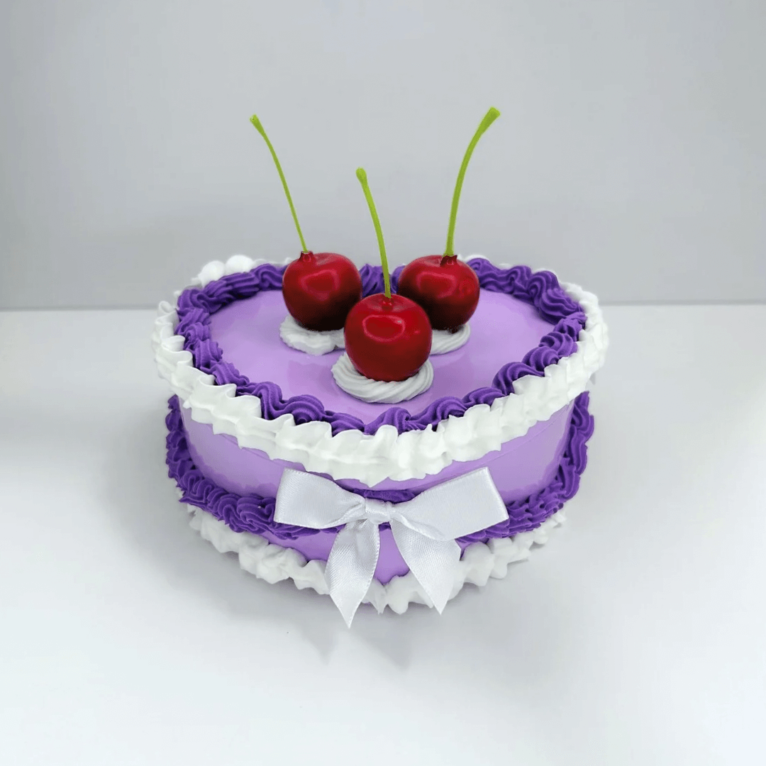 Front view of a charming vintage heart-shaped jewelry box featuring purple and white cream, with three cherries and a white bow on top.