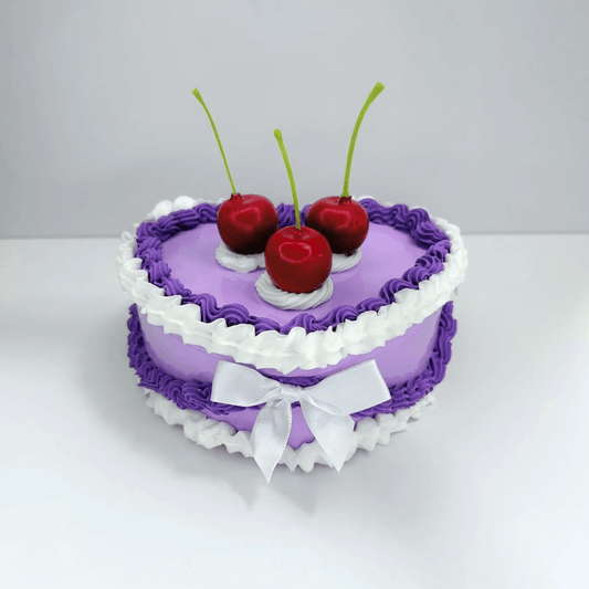 Front view of a charming vintage heart-shaped jewelry box featuring purple and white cream, with three cherries and a white bow on top.