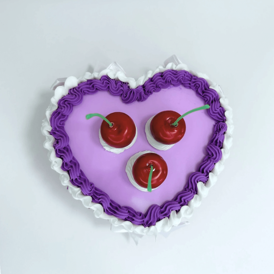 Top view of a vintage heart-shaped jewelry box in purple and white cream colors, topped with three cherries and a delicate white bow.