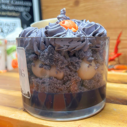 A layered Halloween candle in a clear glass cup, featuring grey frosting, cookie crumbs, white skulls, and a small orange pumpkin decoration on top, set on a wooden surface.