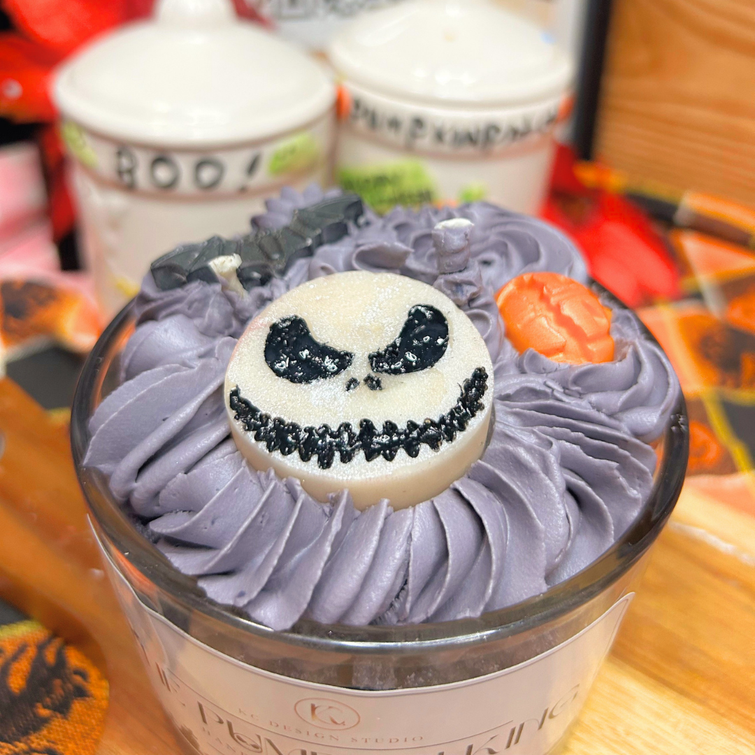 A Halloween-themed candle in a clear glass jar with grey whipped wax, featuring a Jack Skellington face on top, a small orange pumpkin accent, and a black bat decoration. The candle is set on a wooden surface with blurred pumpkins in the background.