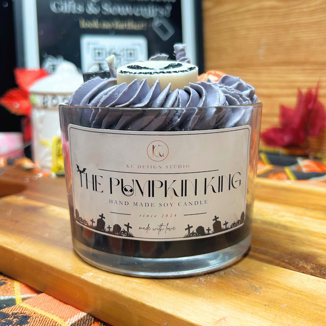 A hand-made soy candle in a clear glass jar with a decorative label reading 'THE PUMPKIN KING' and 'KC DESIGN STUDIO'. The candle has a swirled lavender-gray wax top and a small pumpkin topper, set on a wooden surface with a blurred background.