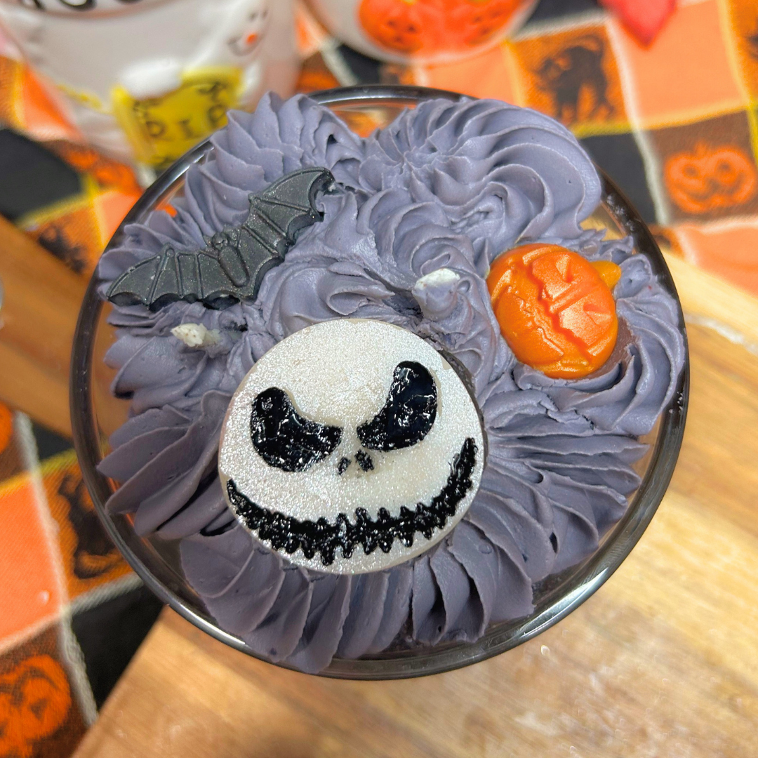 A Halloween-themed candle in a clear glass cup, featuring grey whipped frosting, a Jack Skellington cookie on top, a small bat decoration, and an orange pumpkin topper, set on a wooden table with a patterned tablecloth.