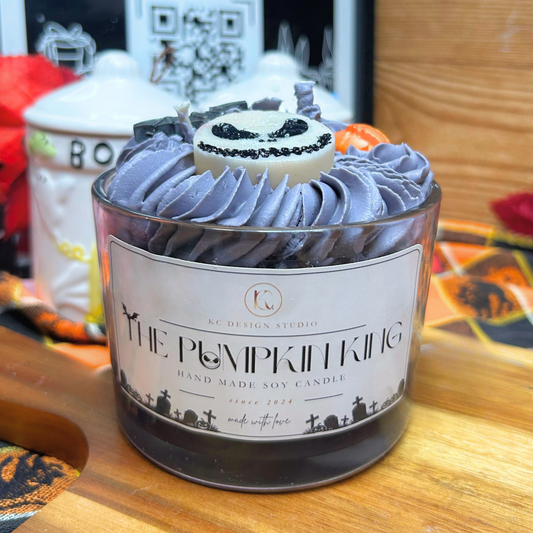 A Halloween-themed soy candle in a clear glass jar with a white label reading 'The Pumpkin King' by KC Design Studio. The candle is topped with gray and black swirled wax resembling frosting, a small white candle with a black pumpkin face, and orange decorative accents, set on a wooden surface.