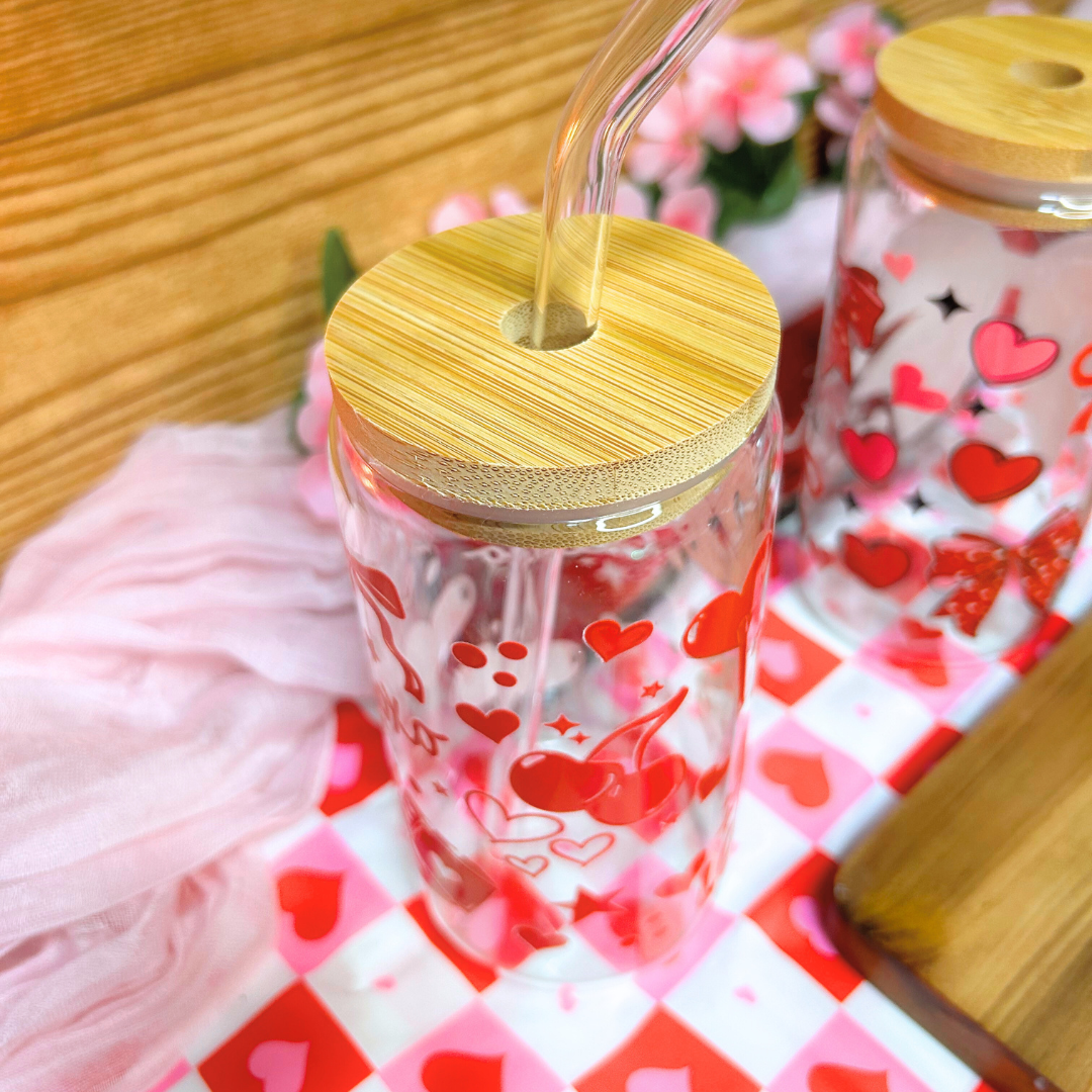 Clear tumbler with red heart designs and a wooden lid on a checkered tablecloth.
